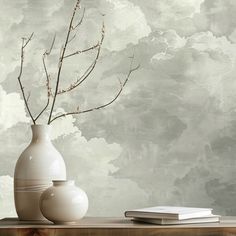 two vases and a book on a table with clouds in the sky behind them