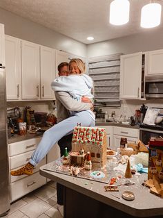 a man and woman hug in the middle of a kitchen with gingerbread houses on the counter