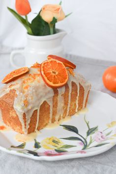 an orange cake with icing on a plate next to some flowers and two oranges