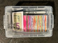 a plastic container filled with lots of different types of pens and paper money on top of a table