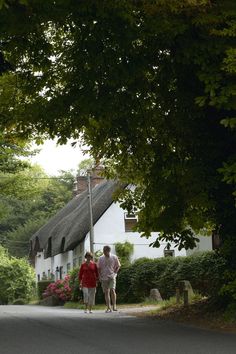 two people walking down the street in front of a white house with thatched roof