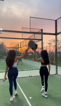 two women holding tennis racquets on top of a tennis court at sunset