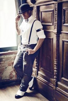 a man leaning against a wall wearing suspenders and a hat