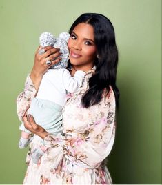 a woman holding a baby in her arms and smiling at the camera while standing against a green wall