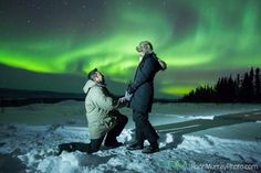 two people standing in the snow with an aurora behind them and one person holding his head