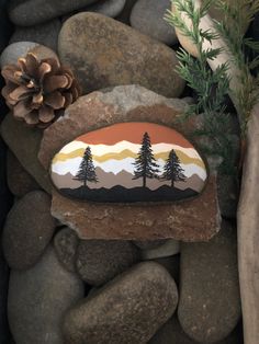 a rock with trees painted on it sitting next to rocks and pine cones in the background