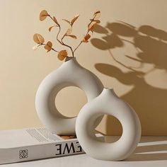 two white vases sitting on top of a table next to a book and plant