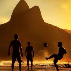 three men playing soccer on the beach at sunset