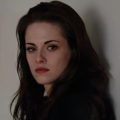 a woman with long dark hair wearing a black jacket and looking at the camera while standing against a white wall