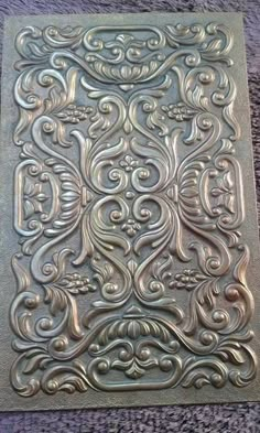 a square metal plate with an intricate design on the front and back side, sitting on a carpeted surface