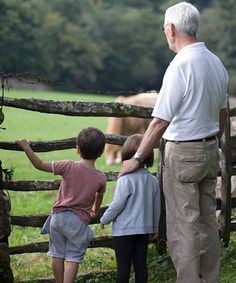 an older man standing next to two young children near a fence with cows in the background