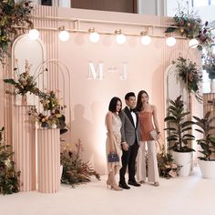 three people standing next to each other in front of a wall with flowers and plants