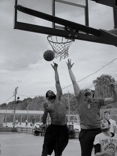 three young men playing basketball on an outdoor court in black and white, with one jumping up to grab the ball