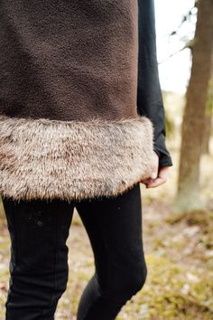 a person wearing black pants and a brown coat with fur on the bottom, standing in front of some trees