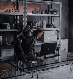 a man sitting in front of a laptop computer on top of a glass table next to a shelf filled with bottles