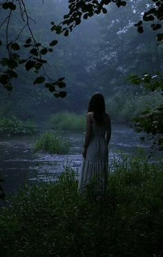 a woman standing in front of a body of water with trees and bushes around her