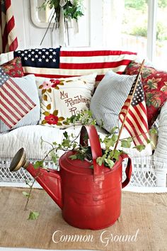 a red watering can filled with flowers on top of a rug next to an american flag pillow