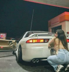 a woman sitting on the ground next to a white car in a parking lot at night