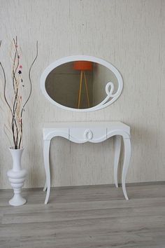 a white table with a mirror and vase on the floor in front of a wall