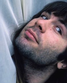 a man with long hair laying on top of a bed next to a white curtain