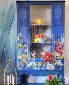 a blue china cabinet with flowers painted on the front and sides, next to a potted plant