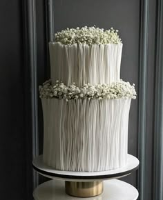 a three tiered cake with white flowers on top is sitting on a stand in front of a gray wall