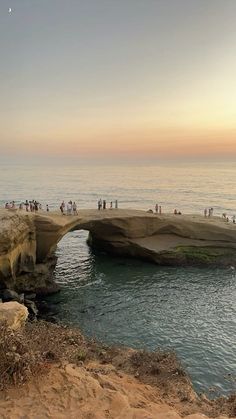 people are standing at the edge of a cliff by the ocean as the sun sets