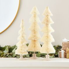 three small white christmas trees sitting on top of a mantle next to presents and a mirror