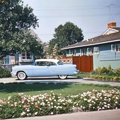 an old blue car parked in front of a house next to a tree and flowers