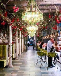 people sitting at tables in a restaurant with chandeliers hanging from the ceiling and flowers on the walls