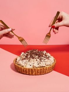 two hands holding forks over a pie on a pink and red surface with one hand reaching for it