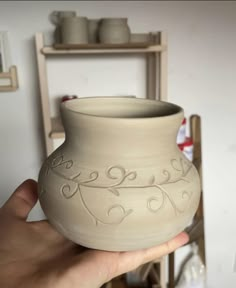 a hand holding a clay vase in front of a shelf