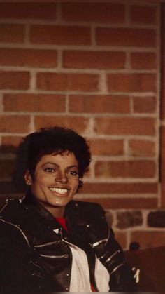 a woman in black leather jacket and red tie sitting next to a brick wall smiling at the camera