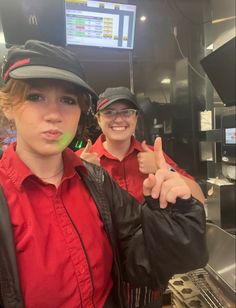 two women in red shirts and black hats posing for the camera with their thumbs up