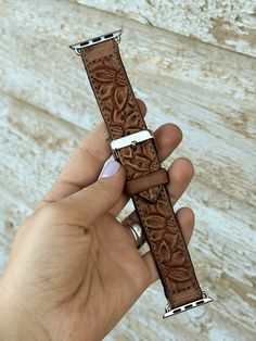 a hand holding a brown watch strap in front of a white brick wall with an intricate design on it