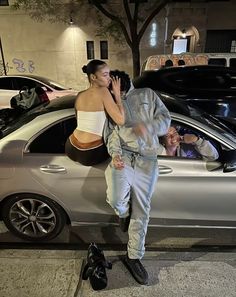 a woman standing next to a silver car talking on a cell phone while another man looks at her