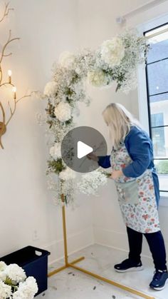 a woman arranging flowers in a room with a gold stand and white roses on the wall