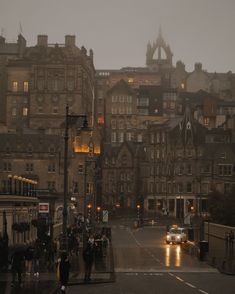 people are walking on the sidewalk in front of some old buildings at night with cars driving down the street