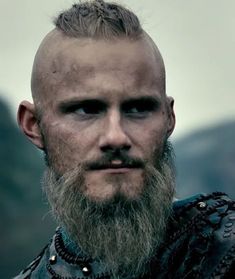 a man with a long beard wearing armor and looking at the camera while standing in front of mountains