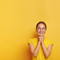 a woman wearing glasses and smiling with her hands folded in front of her face against a yellow background