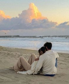 a man and woman sitting on the beach looking out at the ocean under a cloudy sky