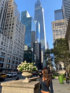 a woman standing in front of tall buildings with flowers on the ledge and people walking down the street