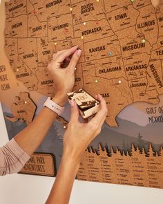 two hands are holding a cell phone in front of a cork board map with the united states on it