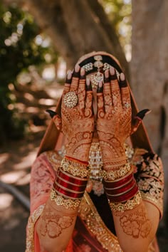 a woman with her hands on her face covered in henna and bracelets, sitting under a tree