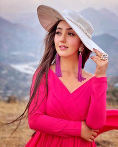 a woman in a pink dress and hat poses for a photo with mountains in the background