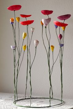 four different colored flowers are in a wire vase on a table with white wall behind them