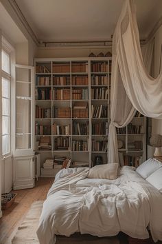 an unmade bed with white sheets and pillows in front of a bookshelf