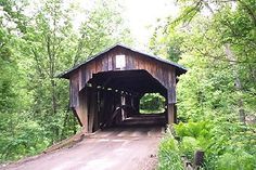 a wooden covered bridge in the woods on a dirt road with lots of trees around it