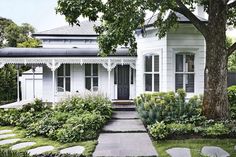 a white house with black shutters and stone walkway leading to the front door is surrounded by greenery