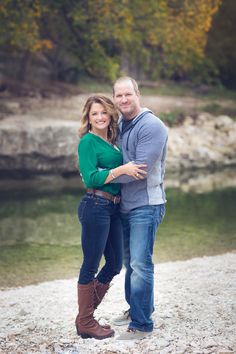 a man and woman standing next to each other in front of a river with trees